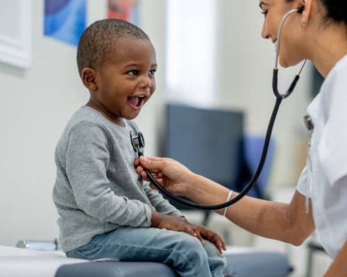 A young boy of African decent, sits up on an exam table as a female Paediatrician preforms a check-up on him.  The boy is dressed casually and smiling as the doctor listens to his heart.